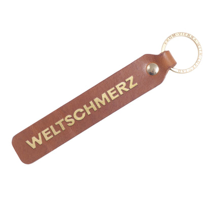 Flatlay, Schlüsselanhänger aus Leder in Natur mit Goldprägung 'Weltschmerz' von LoveTag, handgefertigt in Wien